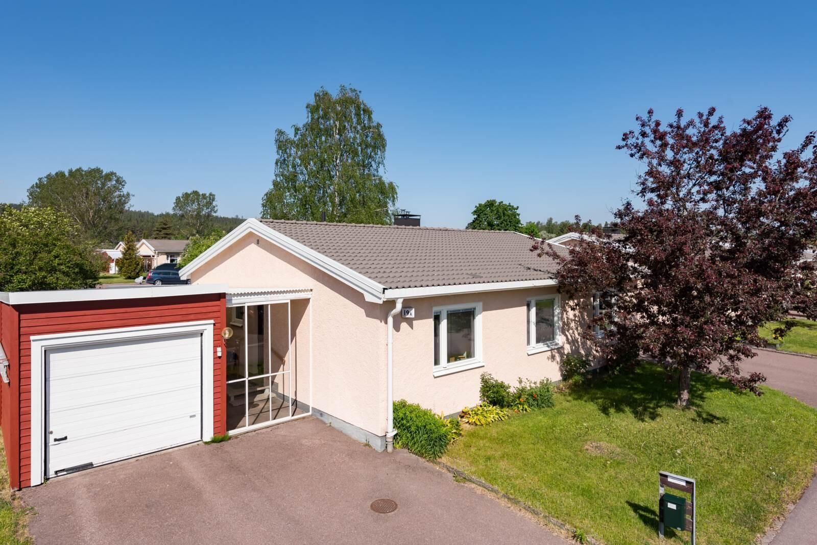 Ljusrosa kedjehus med röd garagelänga, gräsmatta och träd en sommardag.
